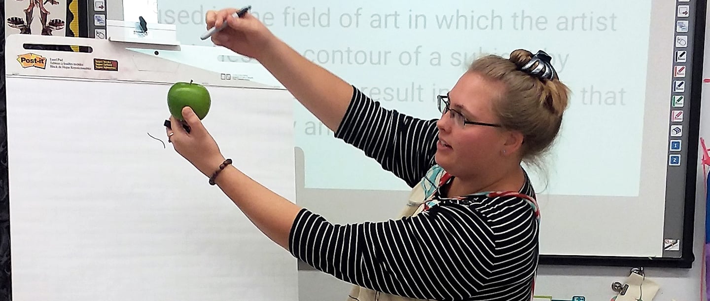 Art instructor holding a green apple in left hand and marker in her right hand t demonstrate sketching