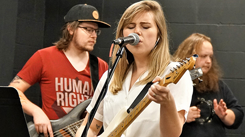a person singing into a microphone with a guitar and a person in the background